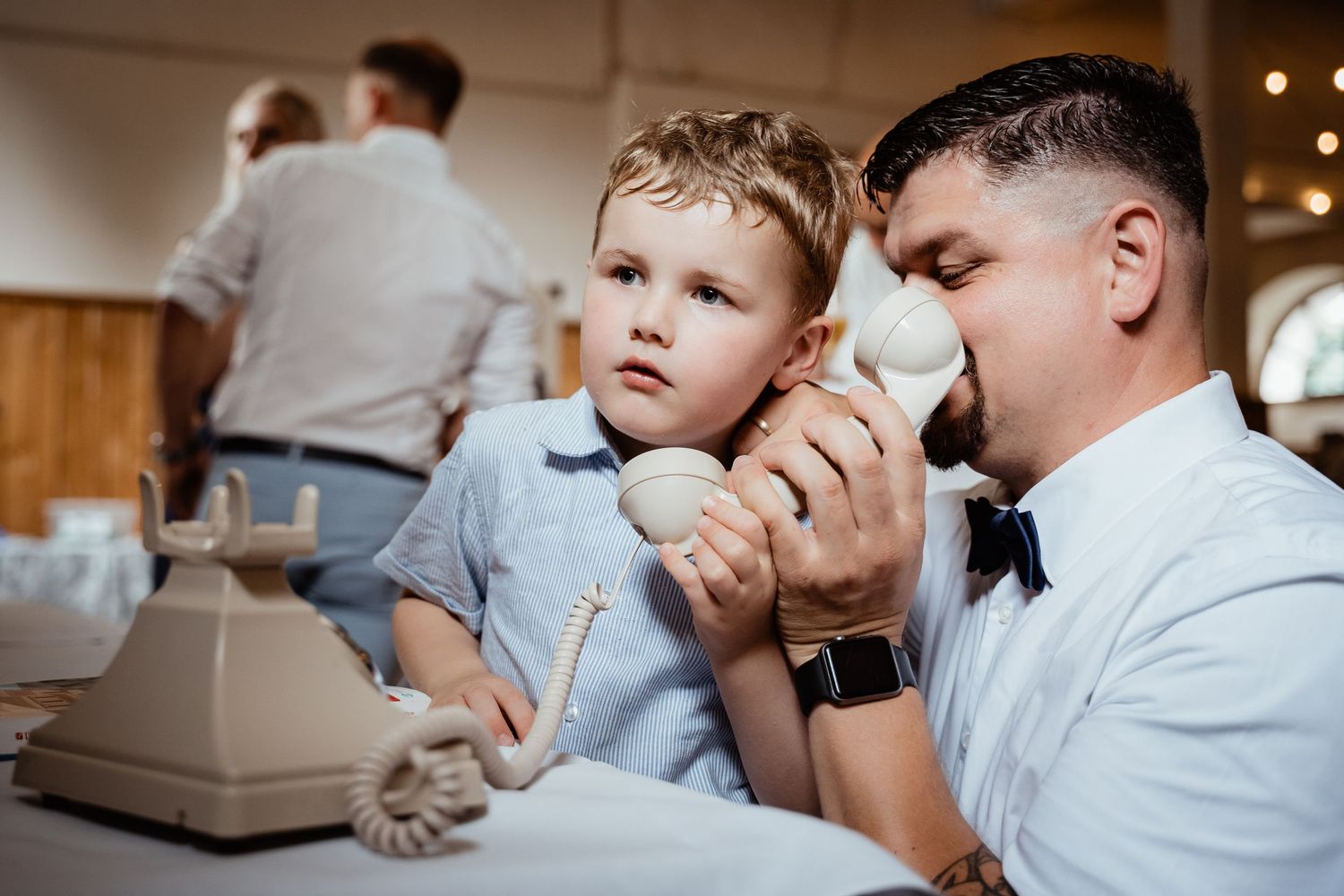 Young boy and man playfully share a vintage rotary telephone handset at a wedding reception table.