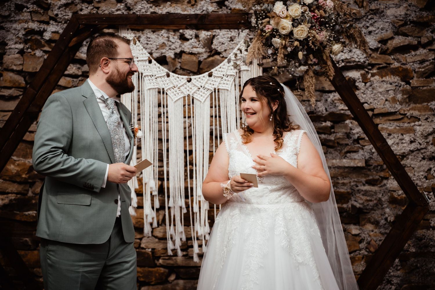 Bride and groom exchanging vows at rustic stone wall altar with macrame wedding backdrop.