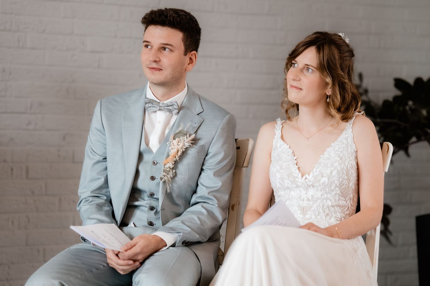 Bride and groom seated together during wedding ceremony, groom in light blue suit, bride in lace dress.
