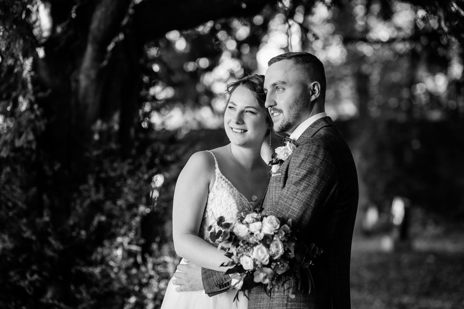 Black and white wedding photo of a smiling bride holding a bouquet and groom embracing outdoors under lush trees.