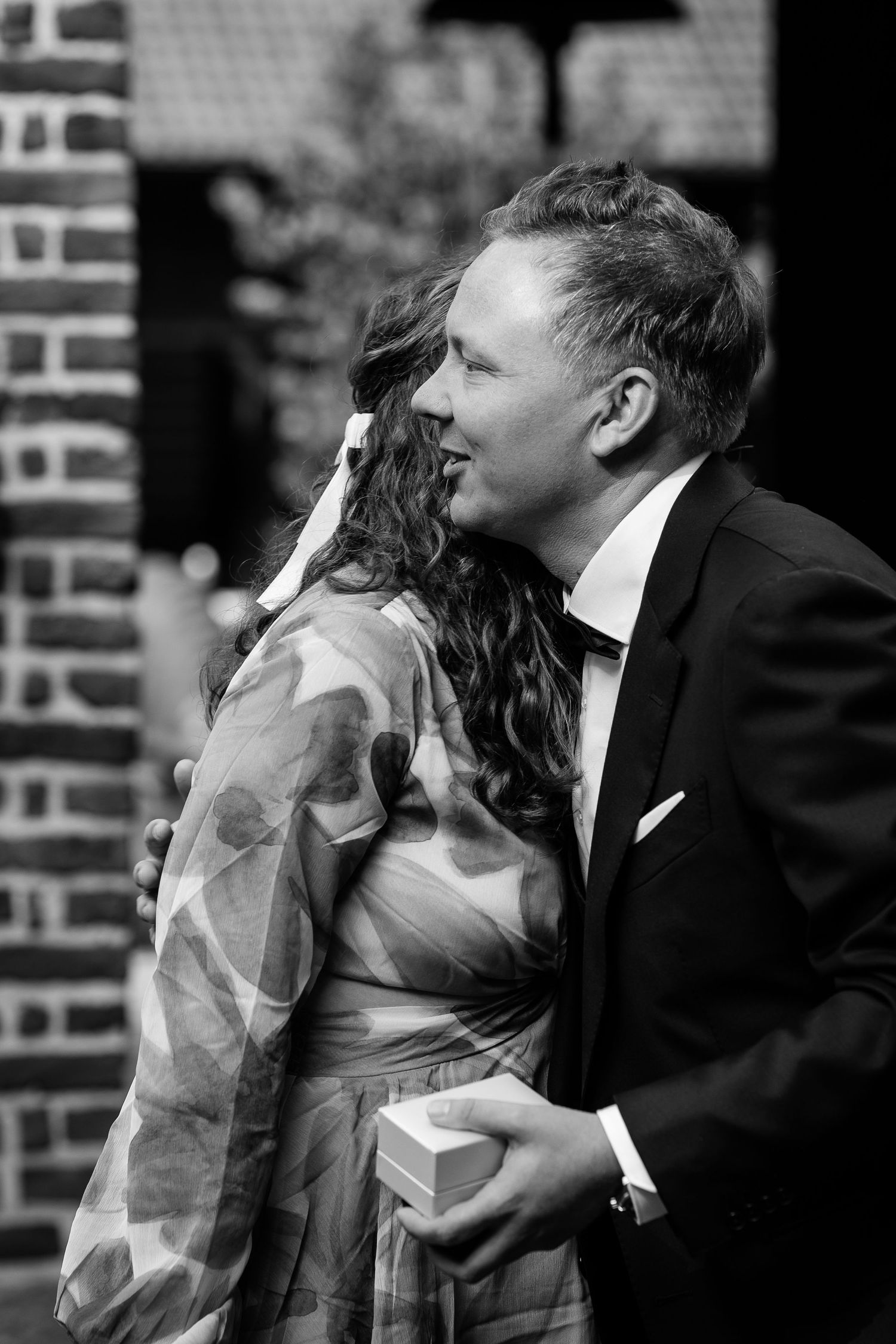 Black and white photo of a couple embracing and smiling outdoors near a brick wall.