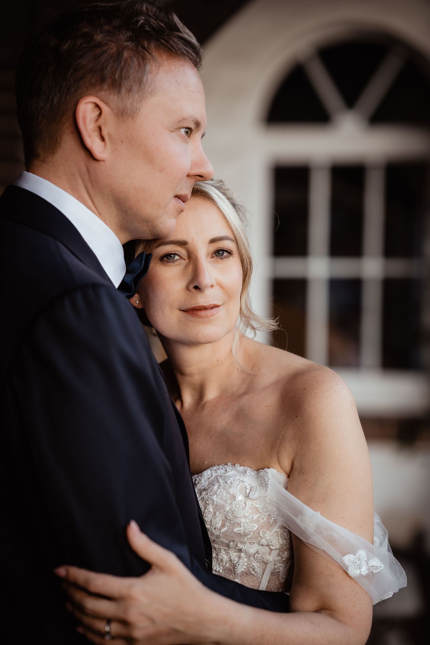 Bride in off-shoulder lace gown embraces groom in dark suit near arched window.