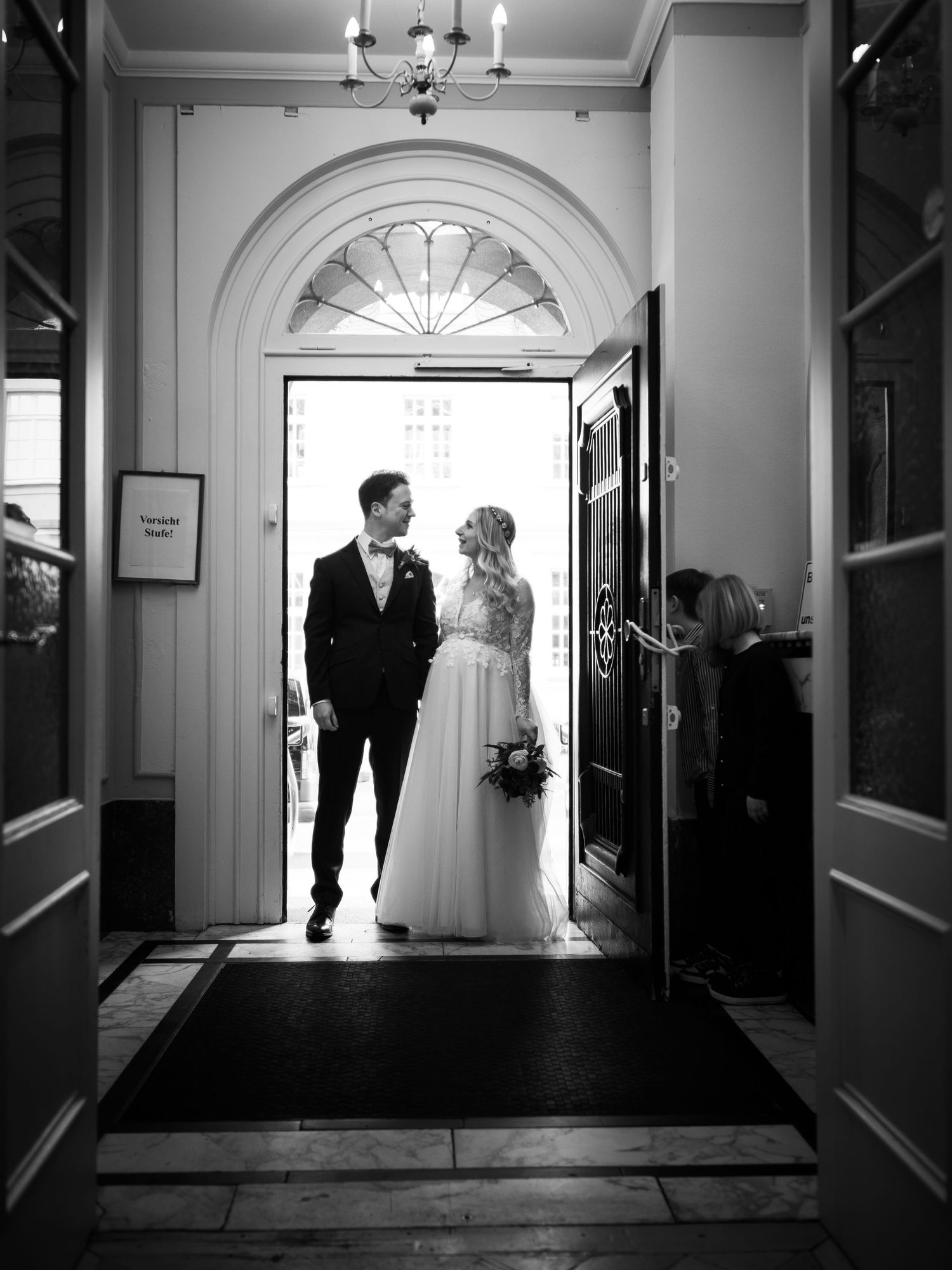 Black and white photo of bride and groom entering an elegant venue through an arched doorway.
