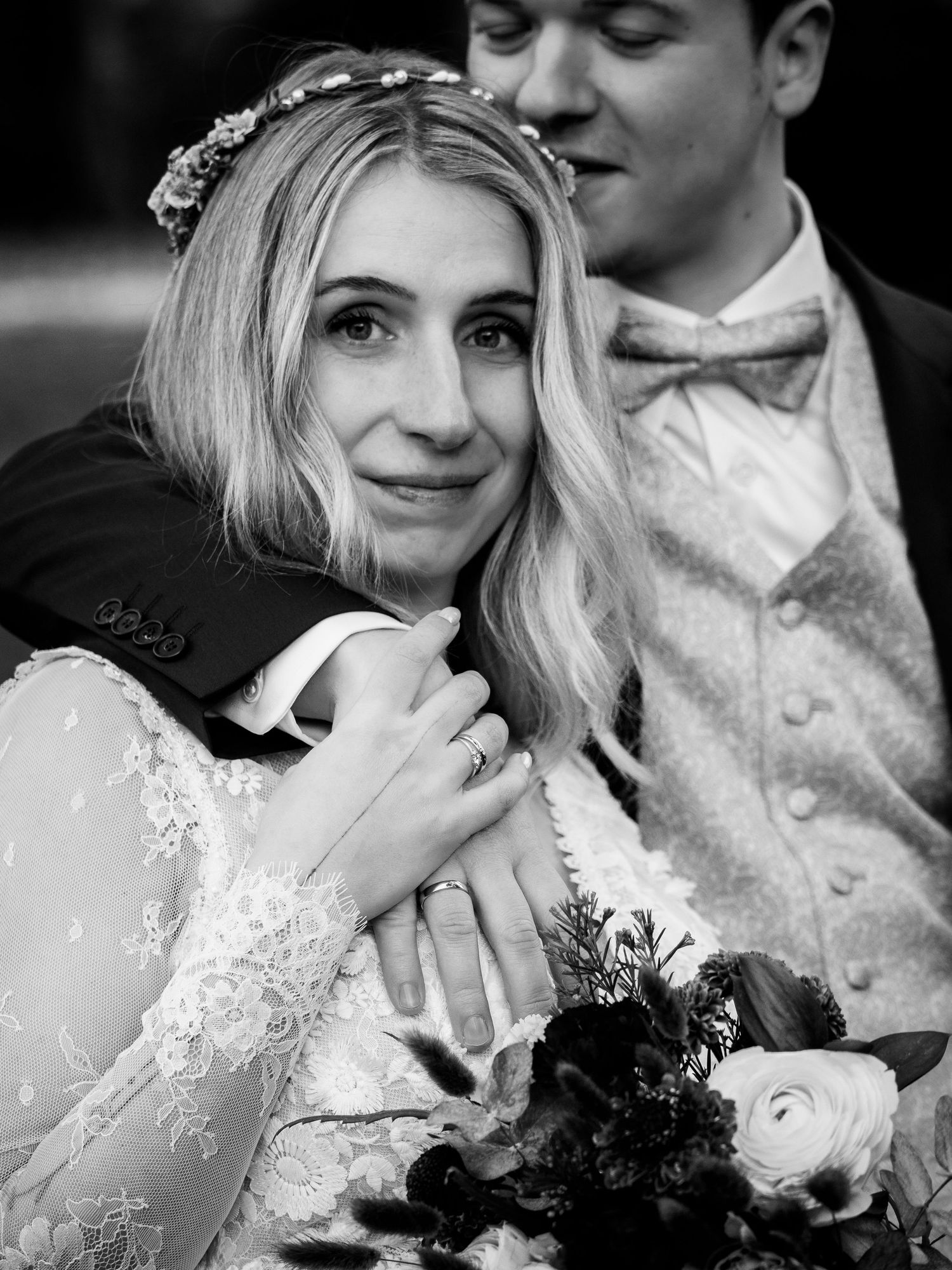 Emotional bride in lace gown holds bouquet while embracing groom in black-and-white wedding portrait.
