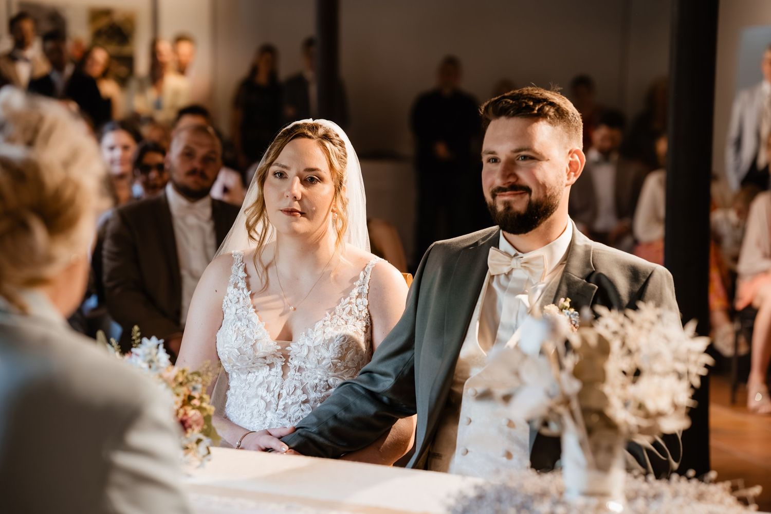 Bride and groom sit together at wedding ceremony altar, smiling, with guests seated in background.