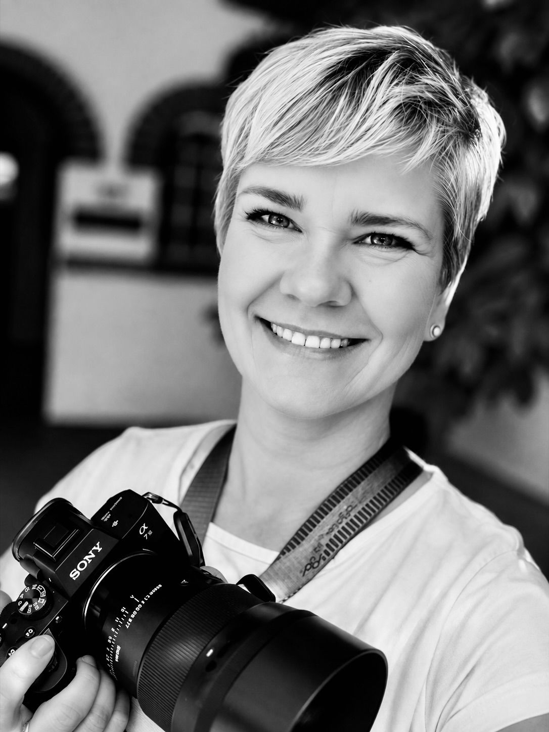 Professional photographer with short blonde hair smiling while holding a camera with a large lens in a black and white portrait.
