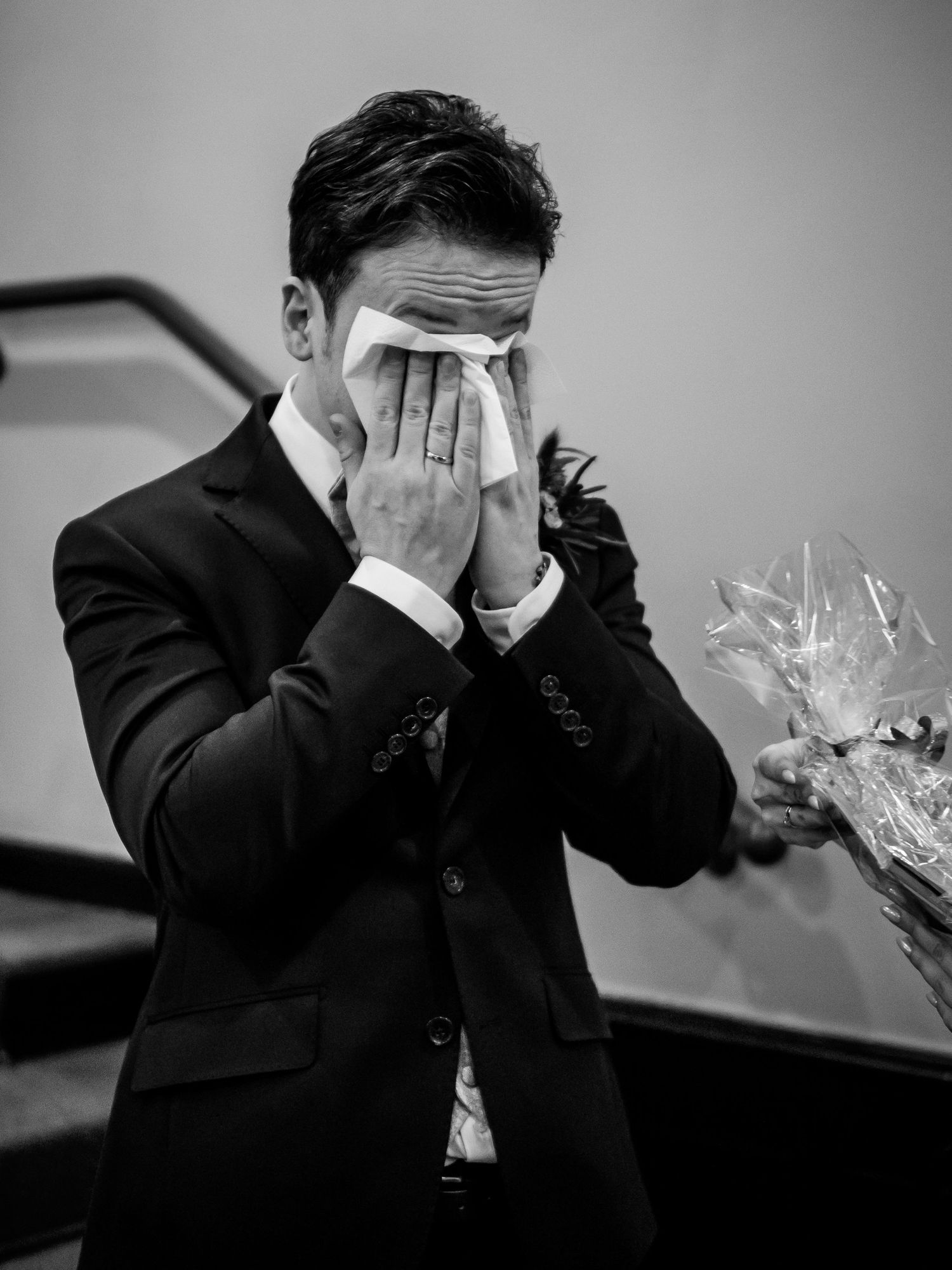Emotional groom in black suit wiping tears with tissue during wedding ceremony in black and white photo.