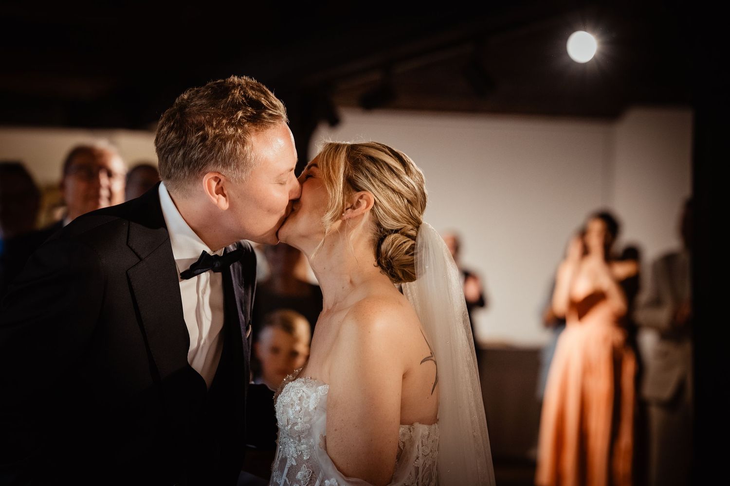 Bride and groom share their first kiss at wedding ceremony, guests applauding in warm amber-lit venue.