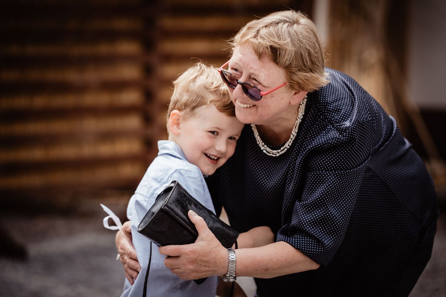 Elderly woman with pearl necklace hugging smiling young boy in light blue shirt outdoors.
