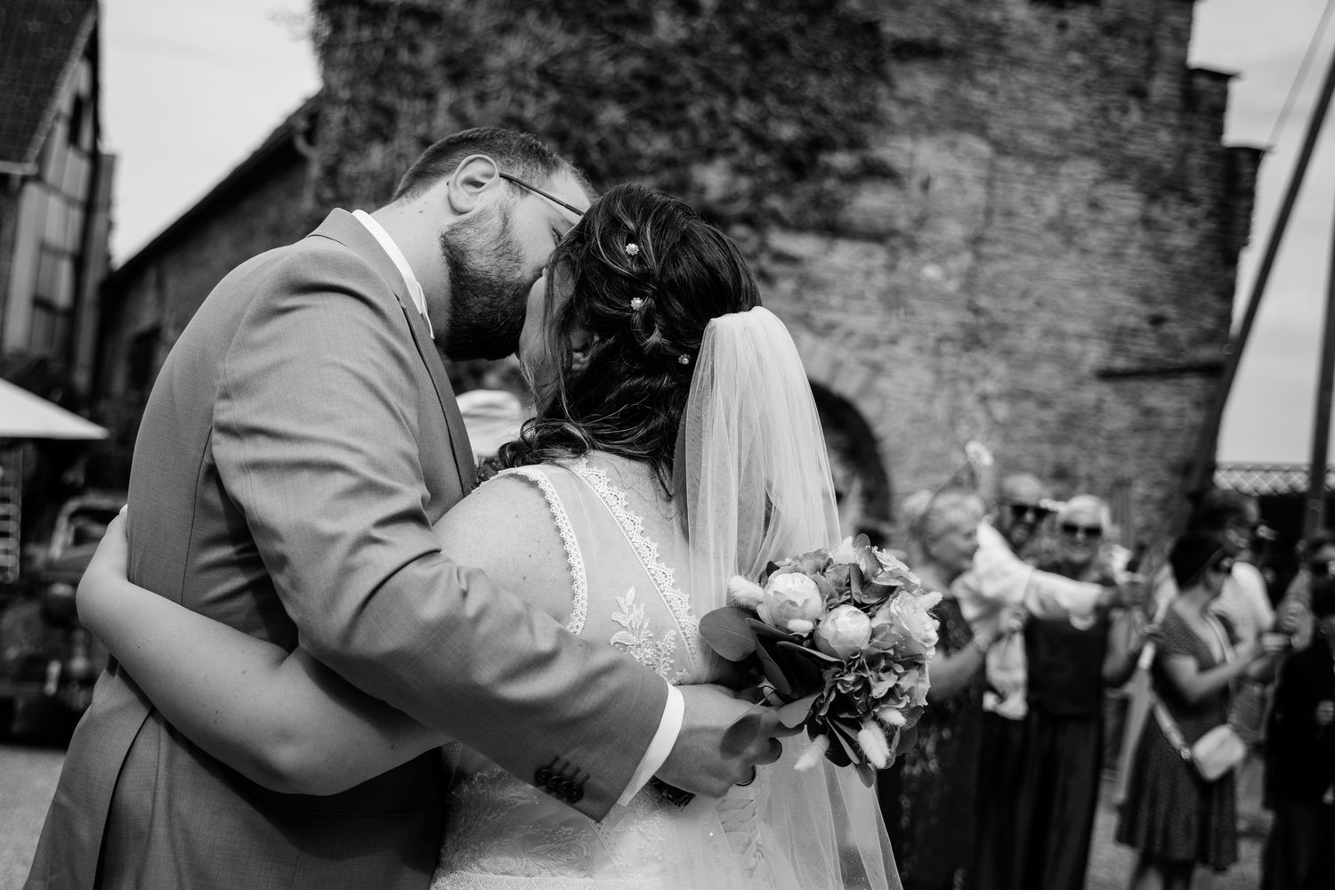 Black and white photo of newlyweds sharing a romantic first kiss outside with guests celebrating.