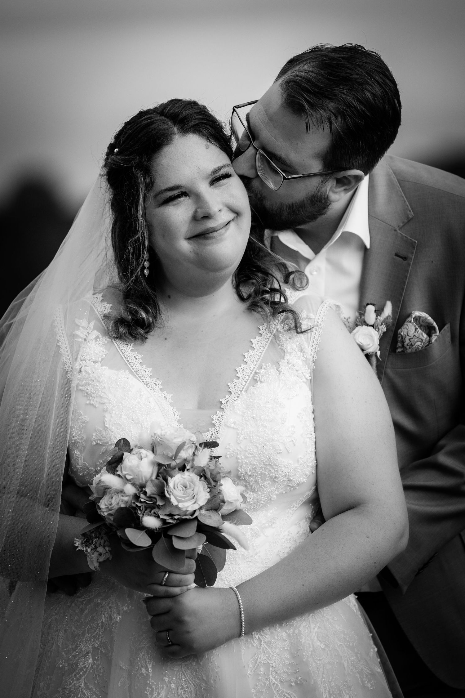 Groom tenderly kissing brides cheek as she smiles holding bouquet in elegant black and white portrait.
