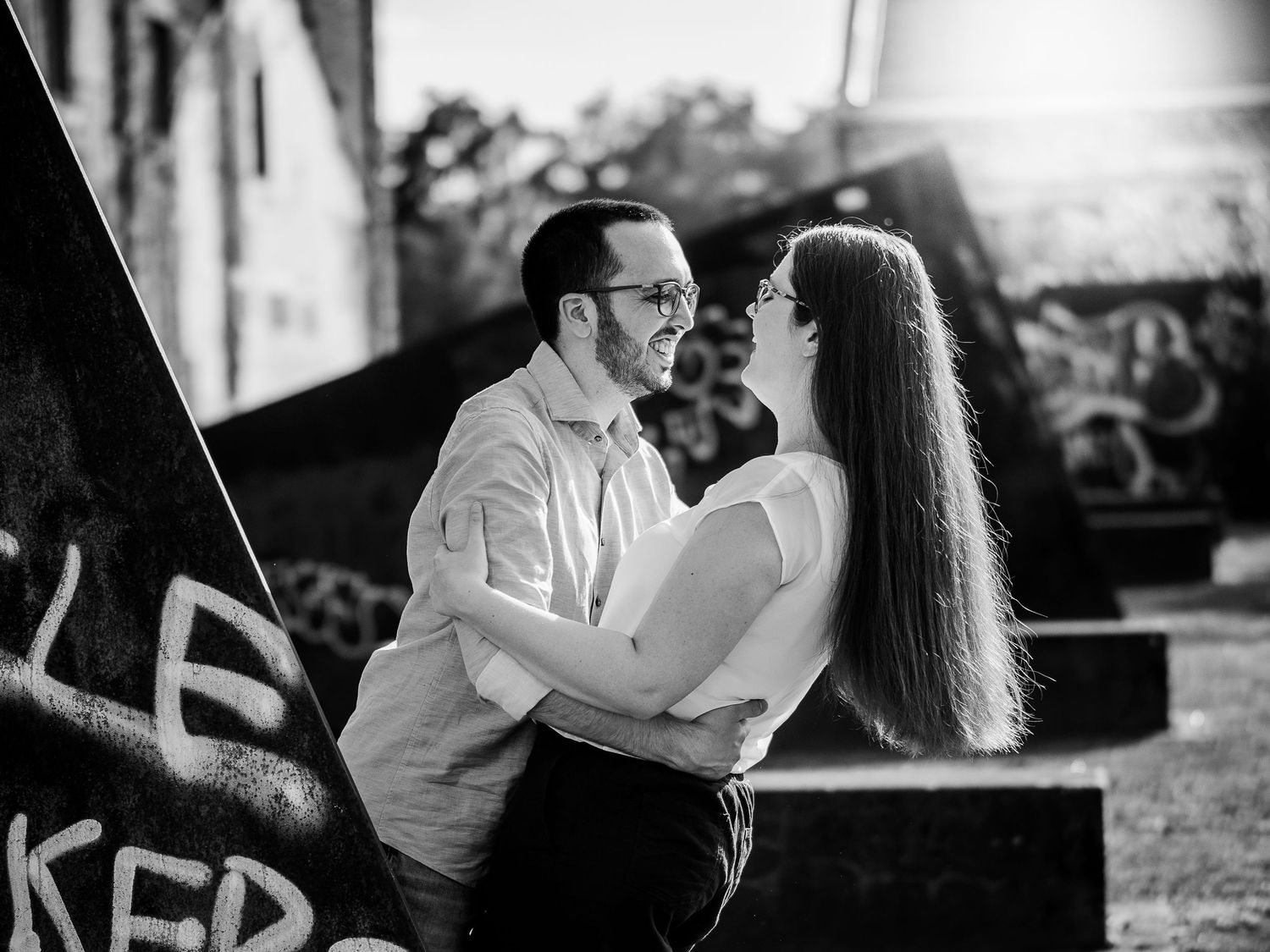 Black and white photo of a couple sharing a romantic moment near graffiti wall outdoors.