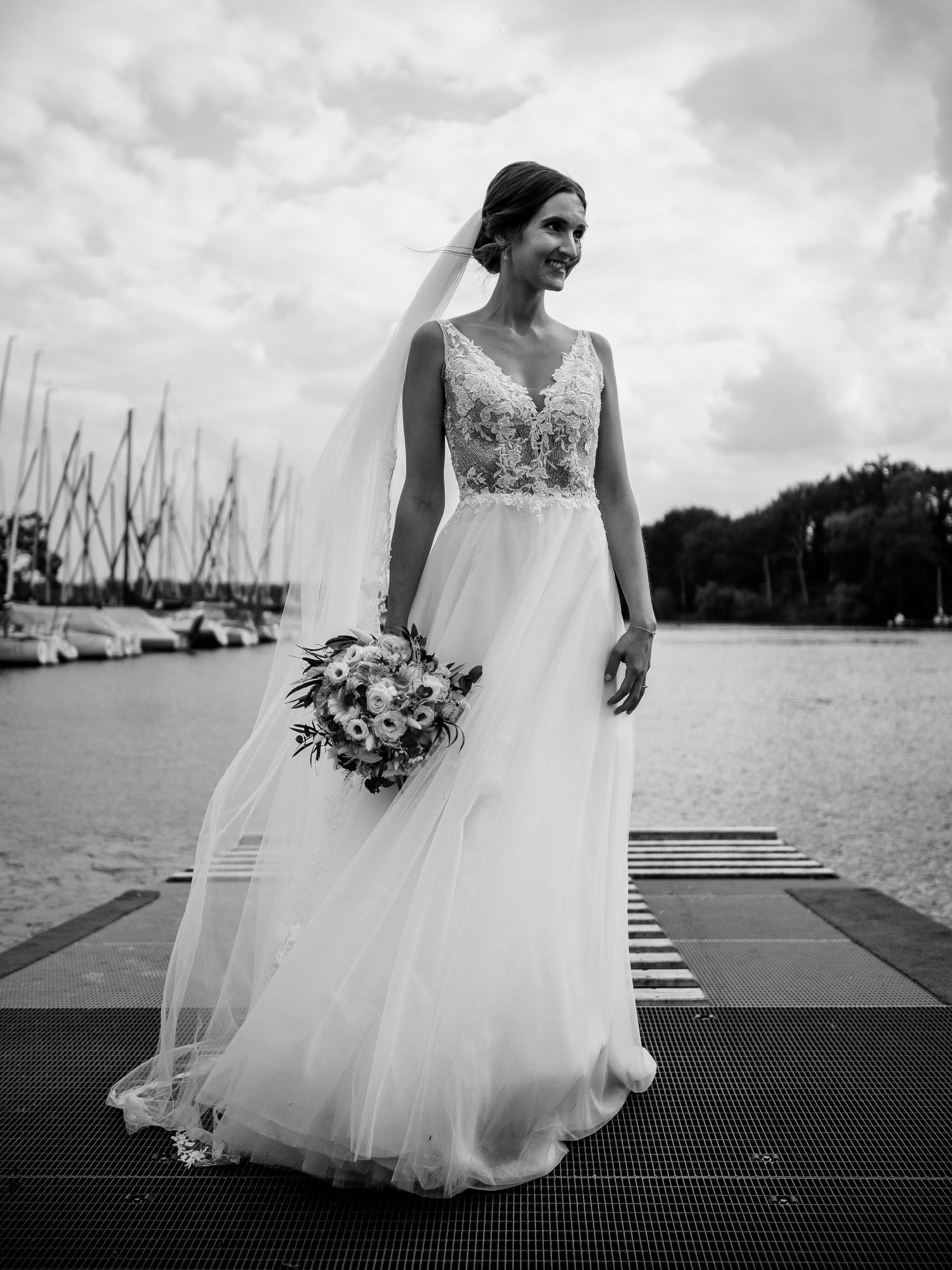 A bride in a flowing white wedding dress stands on a dock near sailboats in a dramatic black and white photo series.