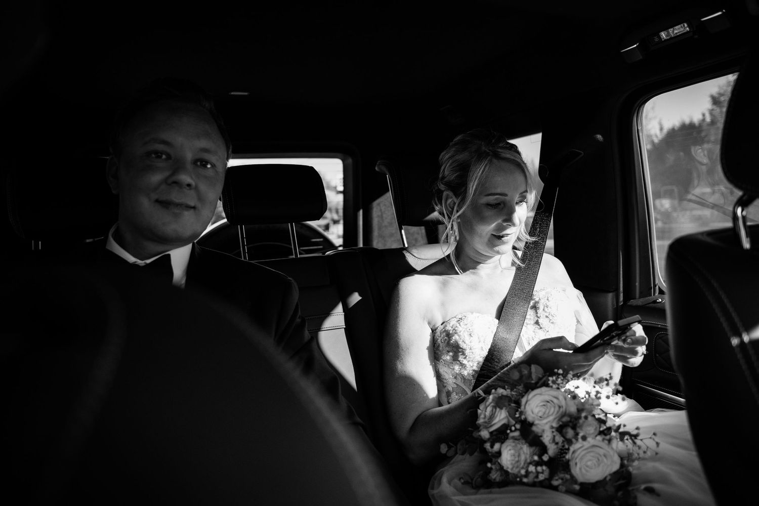 Black and white photo of emotional bride wiping tears in car holding bouquet with man beside her.