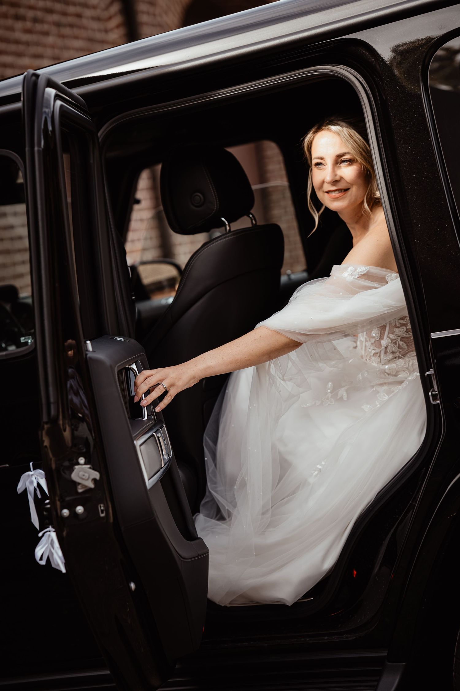 Smiling bride in off-shoulder white tulle gown sitting in black luxury SUV on wedding day.
