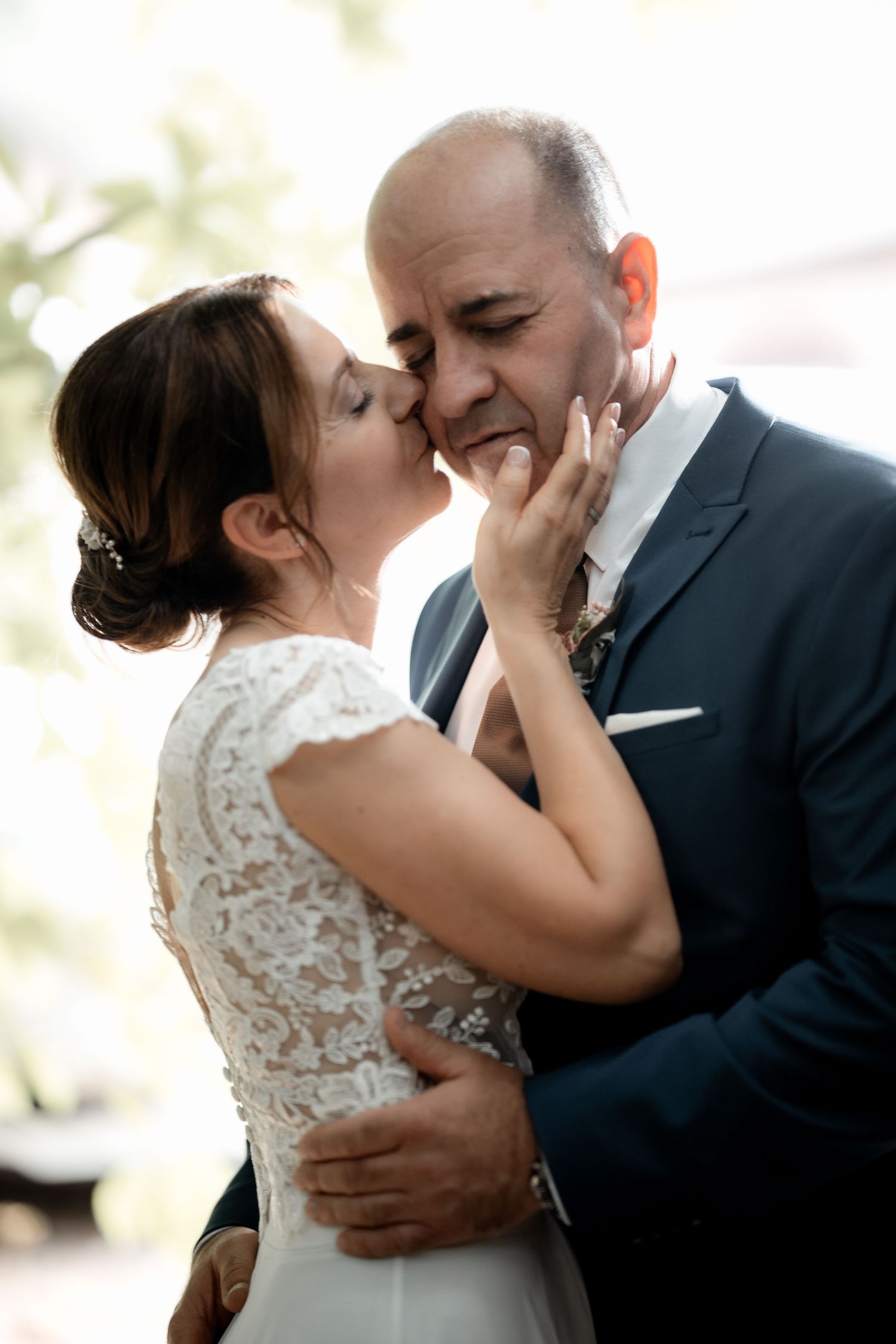 Bride in white lace dress kisses groom tenderly on cheek in romantic close-up wedding portrait.