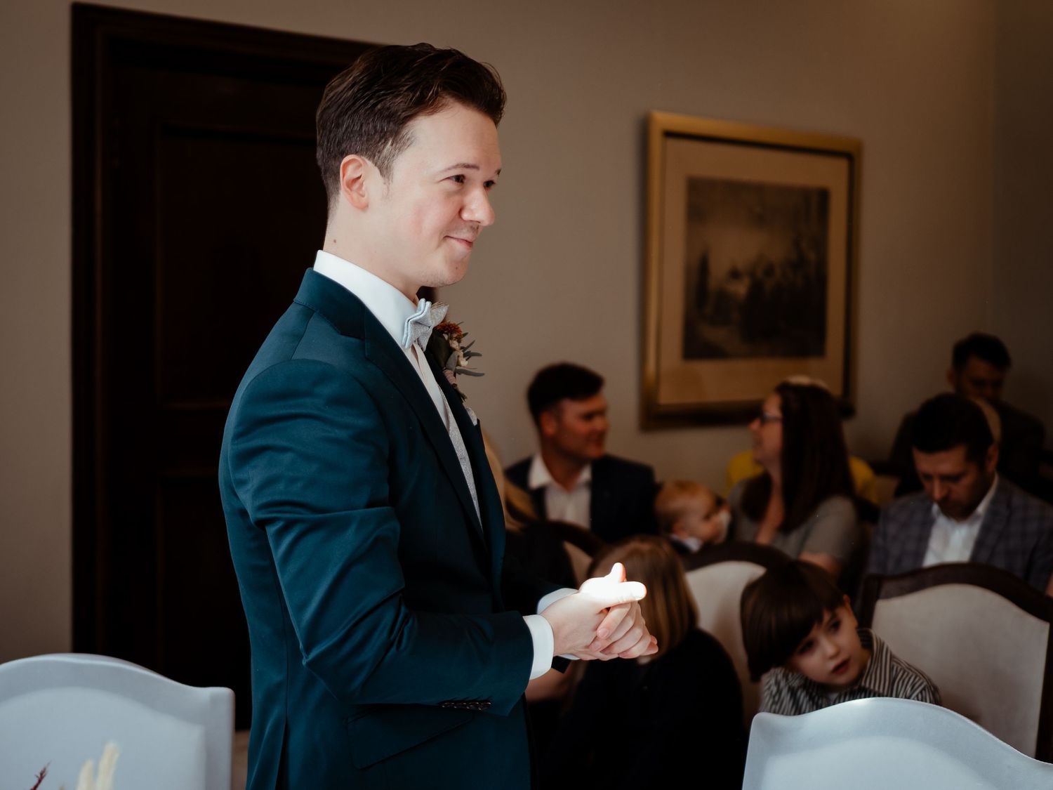 Groom in teal velvet suit holding candle at wedding ceremony with guests seated in background.