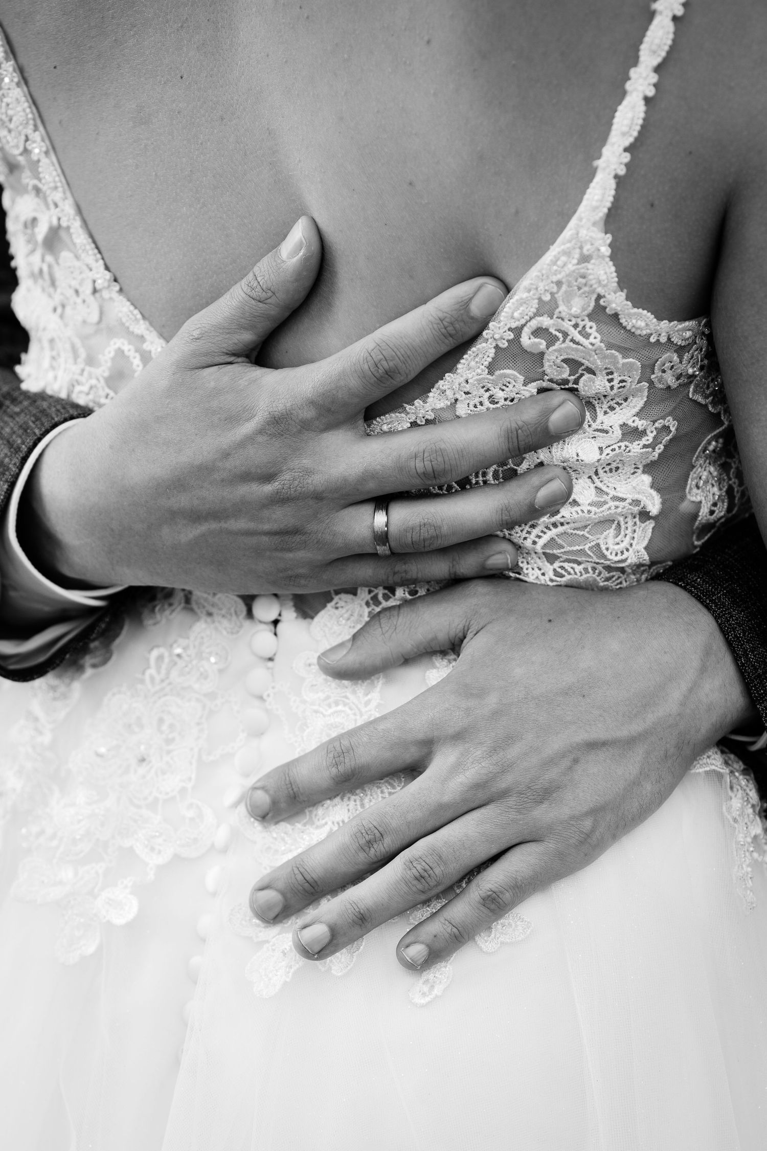 Black and white photo of newlyweds embracing, showing wedding rings on their hands against a lace bridal gown.
