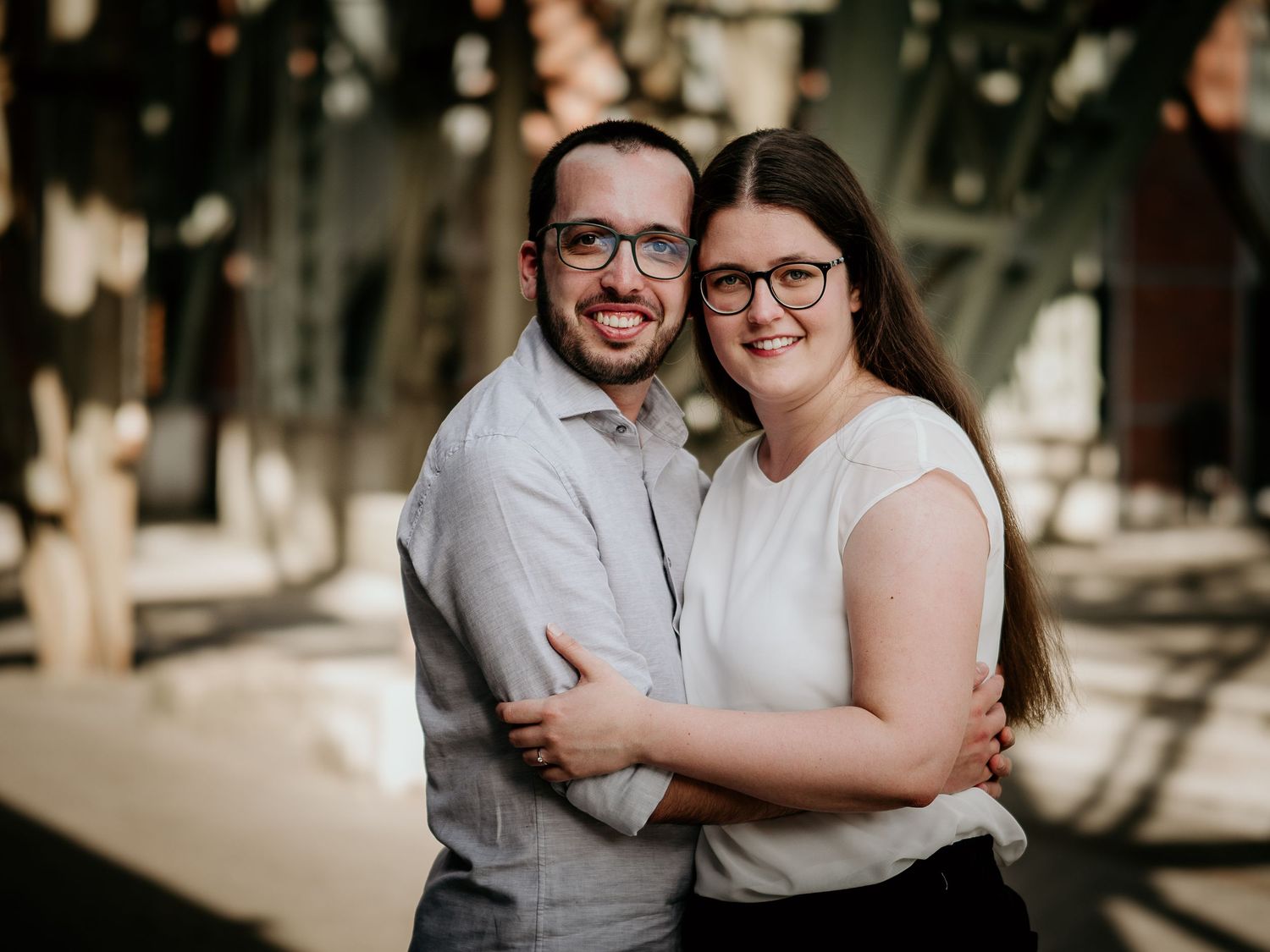 Happy couple in glasses embracing outdoors, smiling warmly in a sunlit urban setting.