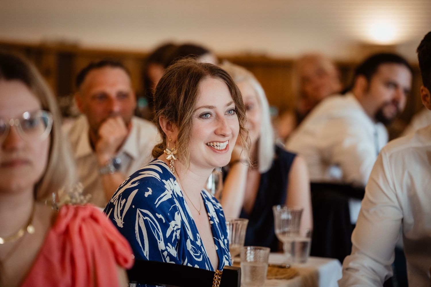 A smiling woman in a blue floral dress enjoys a wedding reception, surrounded by guests in a warmly lit venue.