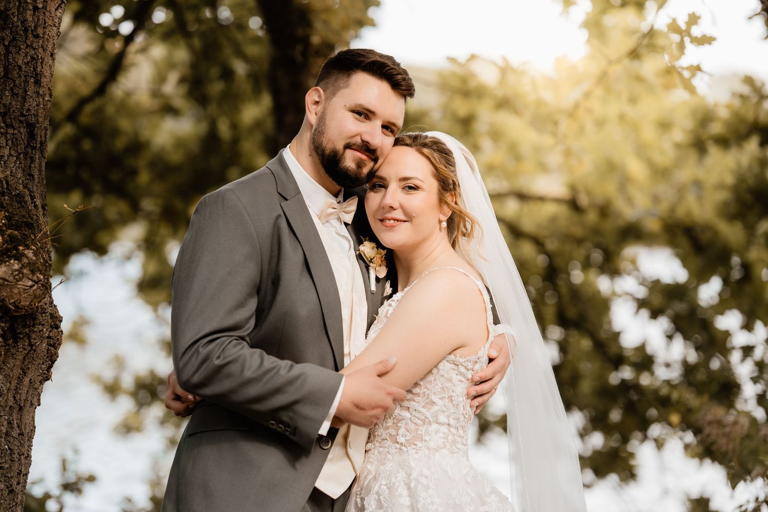 Bride and groom embrace outdoors under green trees, wearing wedding dress with veil and gray suit.