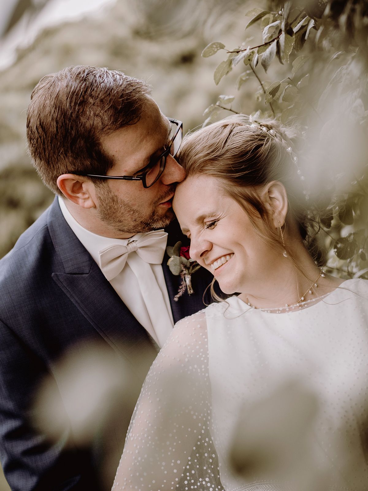 A groom in a navy suit kisses his smiling bride on the forehead outdoors surrounded by lush greenery.