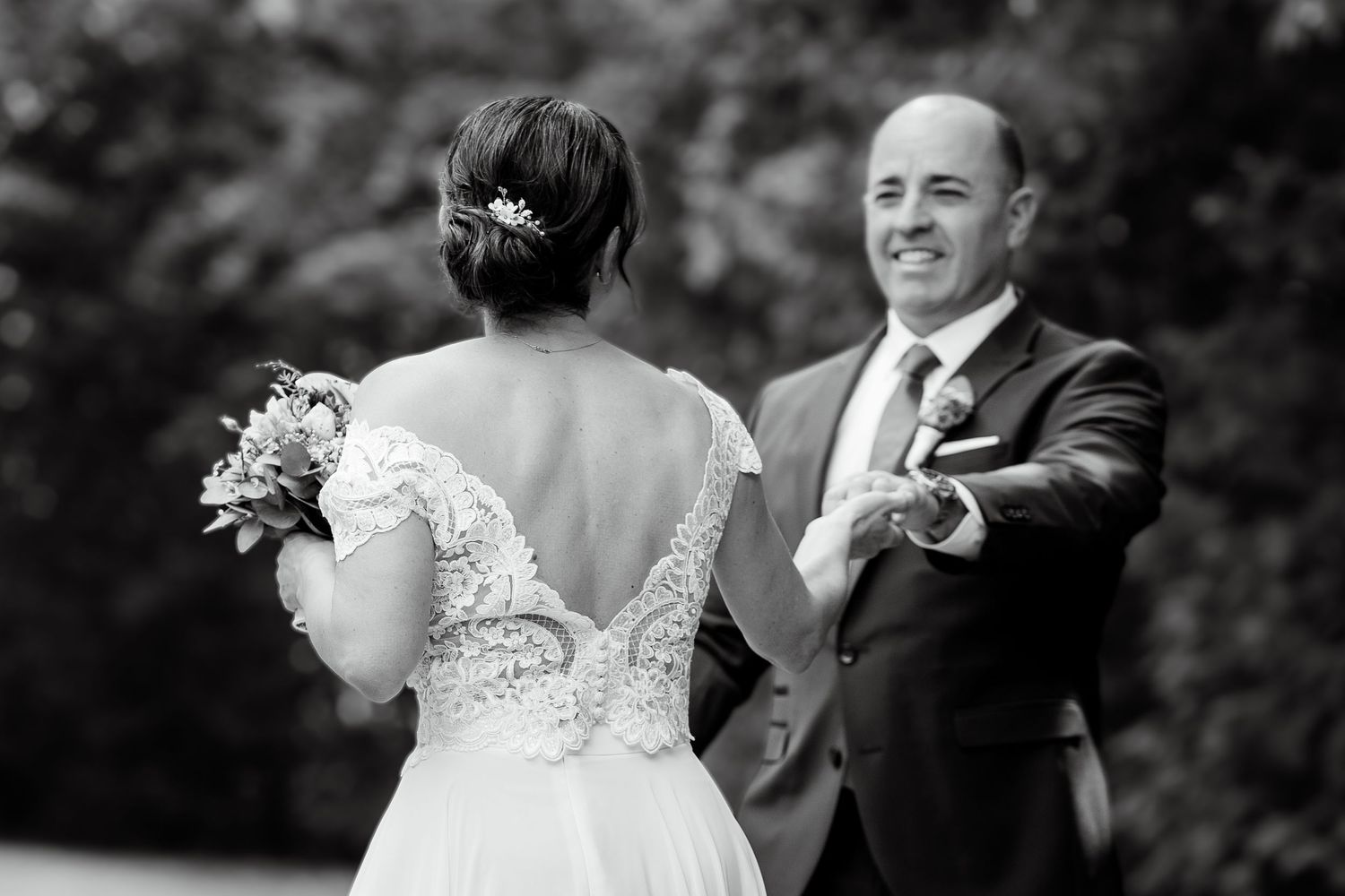 Black and white photo of a bride in lace gown holding bouquet, sharing a joyful moment with a smiling man in a suit.