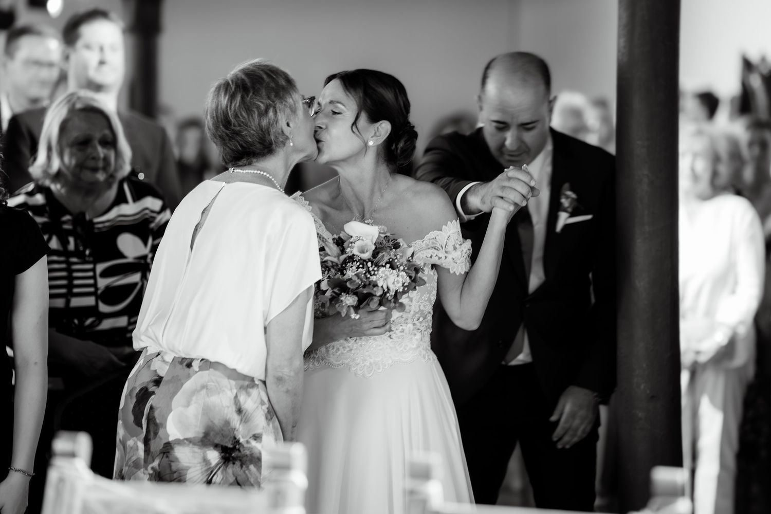 Bride holding bouquet receives a kiss from a guest while groom stands nearby at wedding reception.