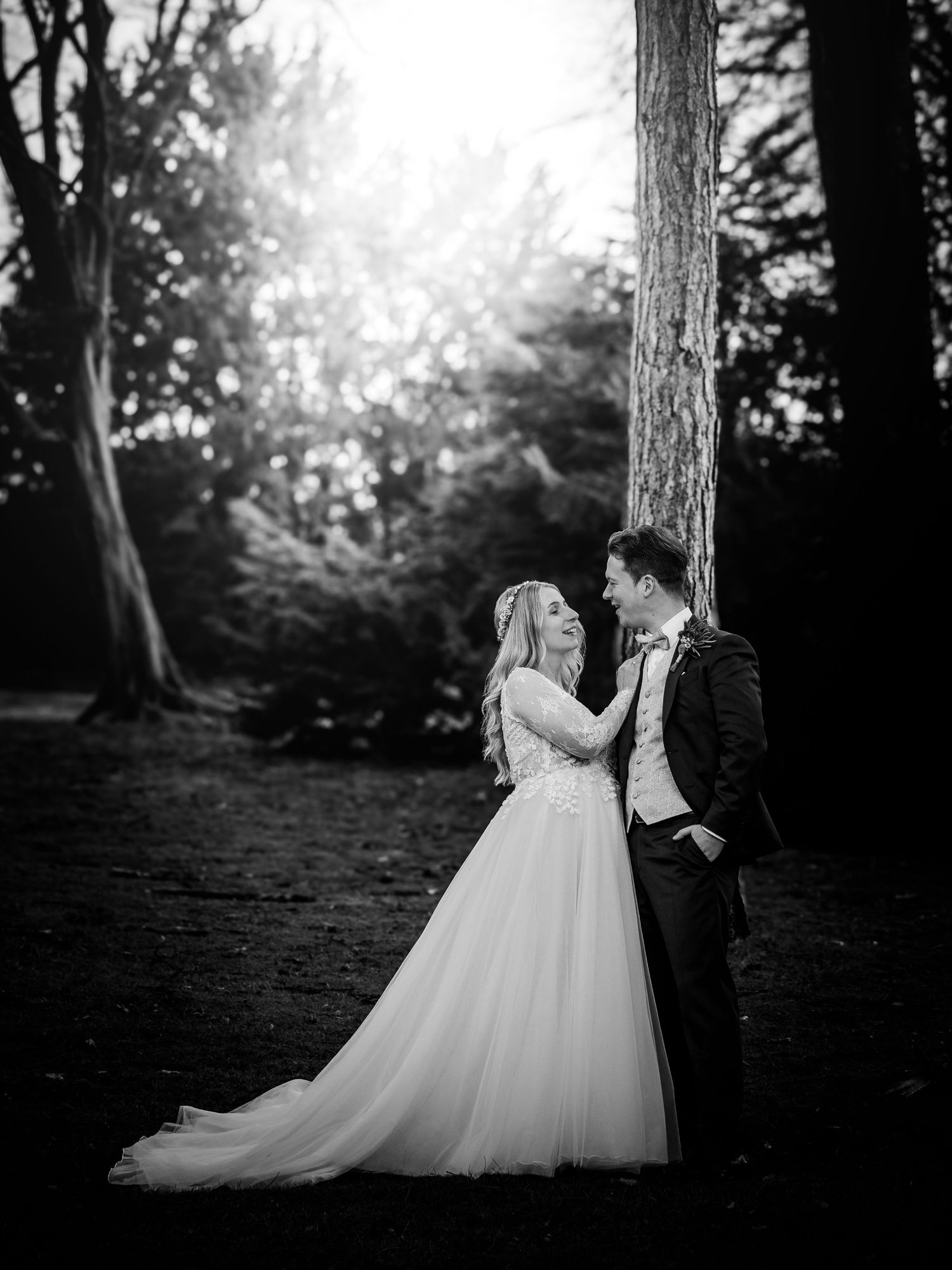 Black and white photo of bride and groom gazing at each other beside a tall tree in woods.