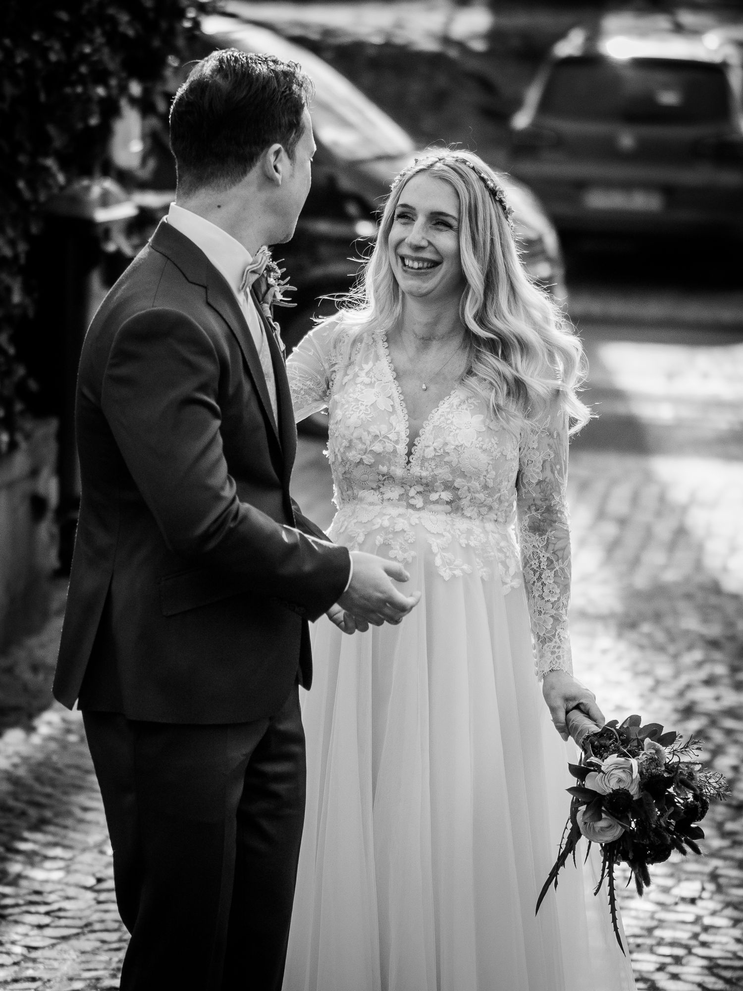 Bride in lace wedding gown smiles at groom in black suit while holding bouquet on cobblestone street.