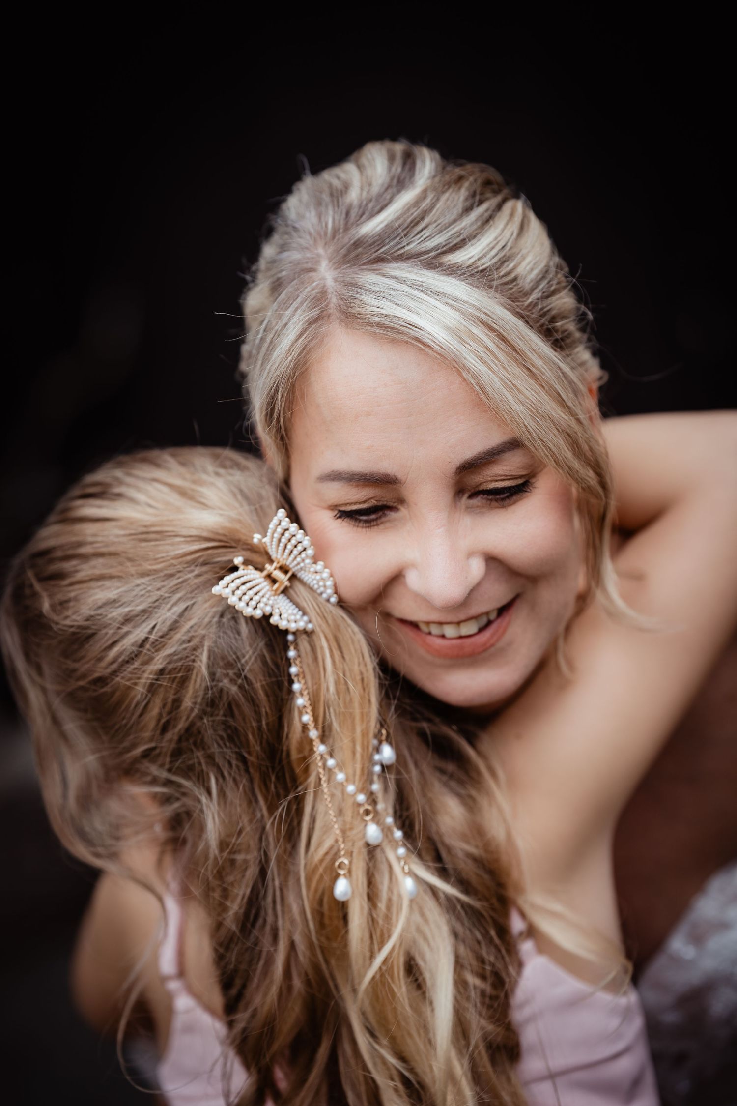 Smiling blonde bridesmaid with butterfly hair accessory sharing a warm embrace at a wedding.