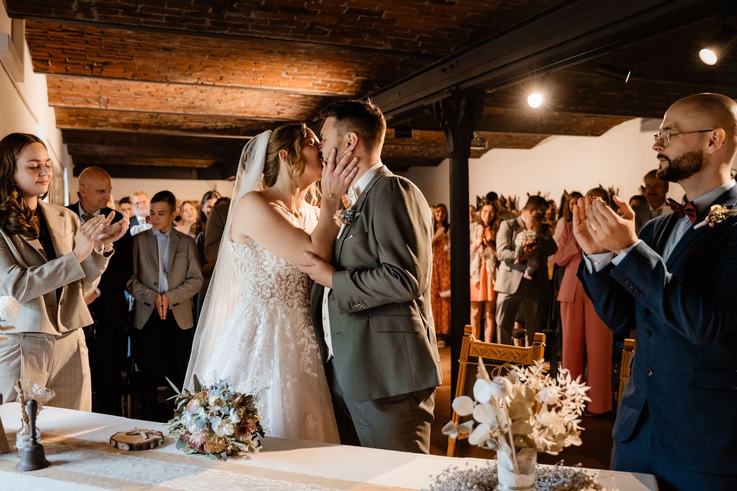 Bride and groom share first kiss at wedding ceremony while guests applaud in a warmly lit rustic venue.