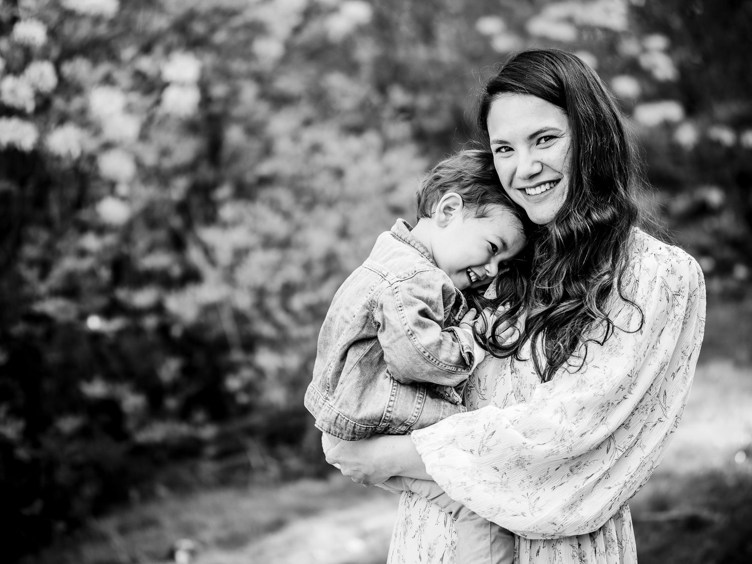 Black and white photo of a smiling mother tenderly hugging her young child outdoors in a garden setting.