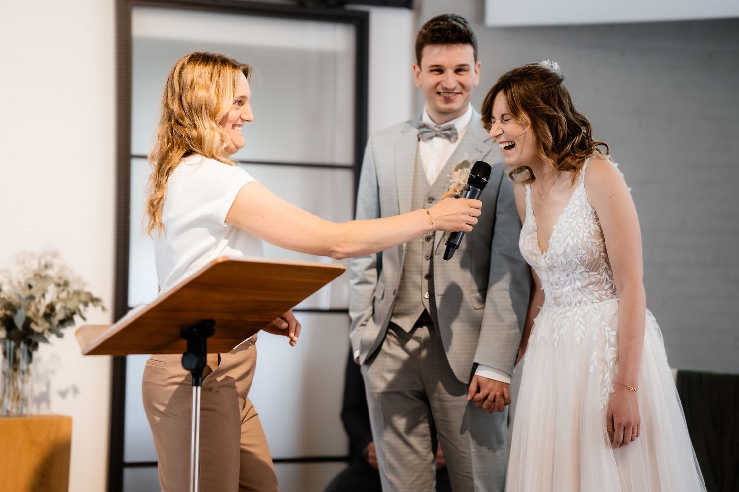 Officiant holds microphone to laughing bride while groom smiles during wedding ceremony.