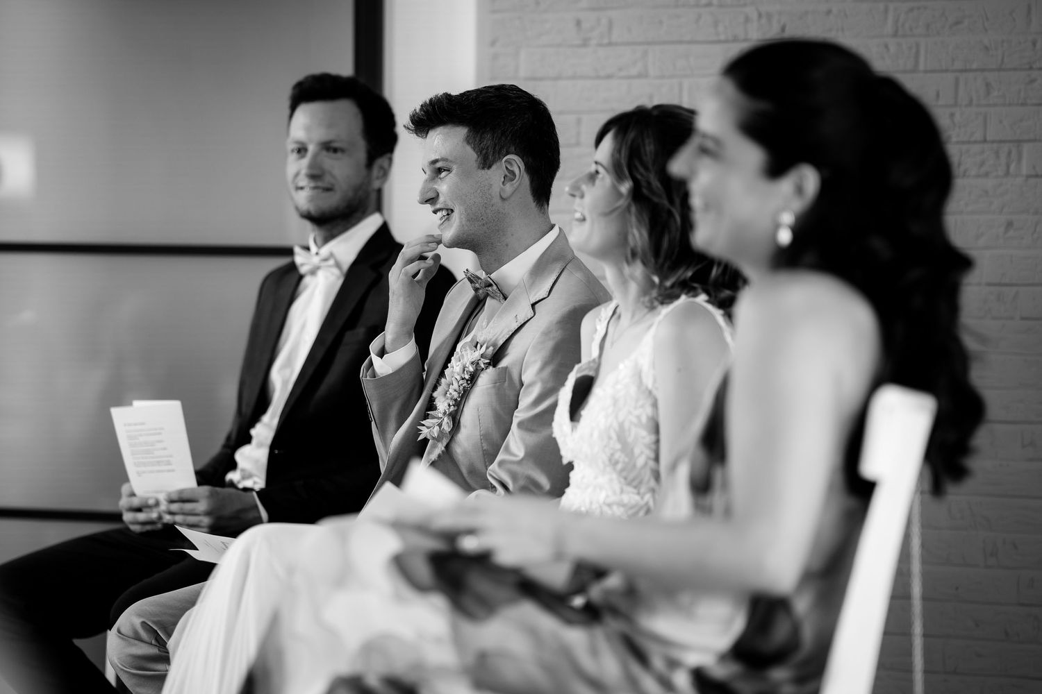 Black and white photo of wedding guests laughing and smiling during a ceremony seated in white chairs.