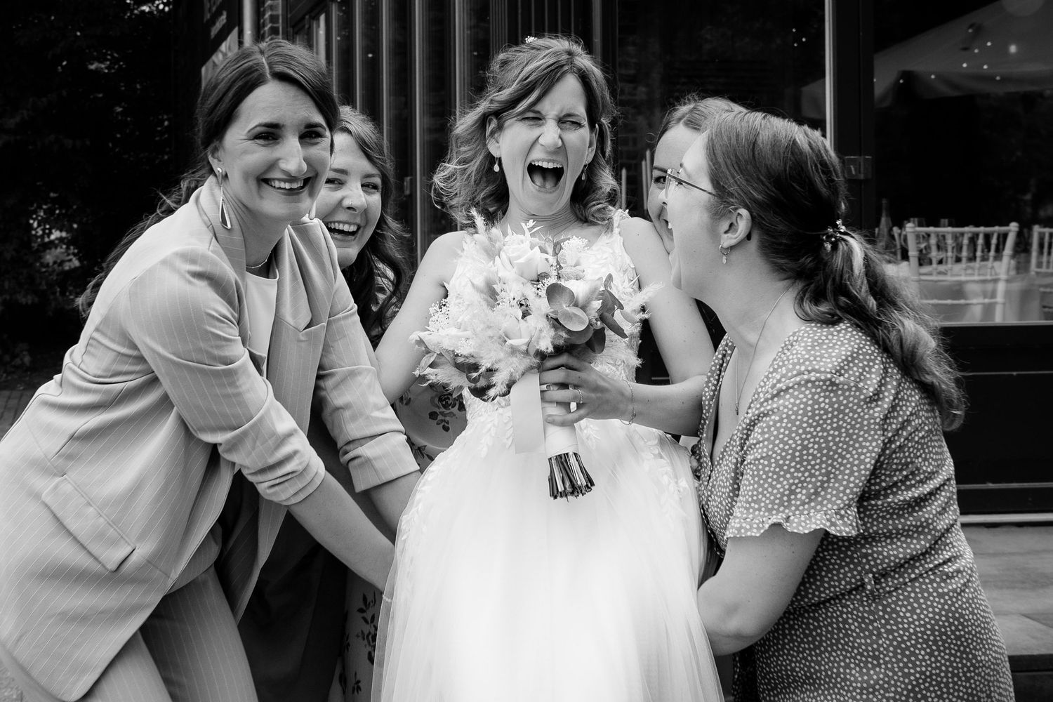 Bride laughing joyfully with bridesmaids while holding a bouquet in black and white photo.