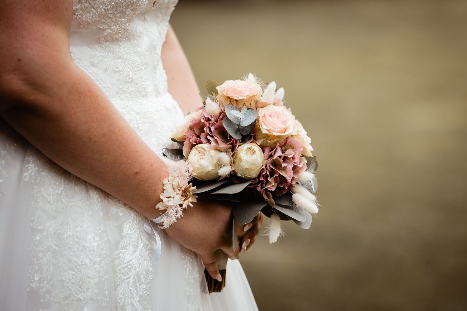 Bride in white lace wedding dress holds a elegant bouquet of pink roses, dried flowers, and pearl accents.