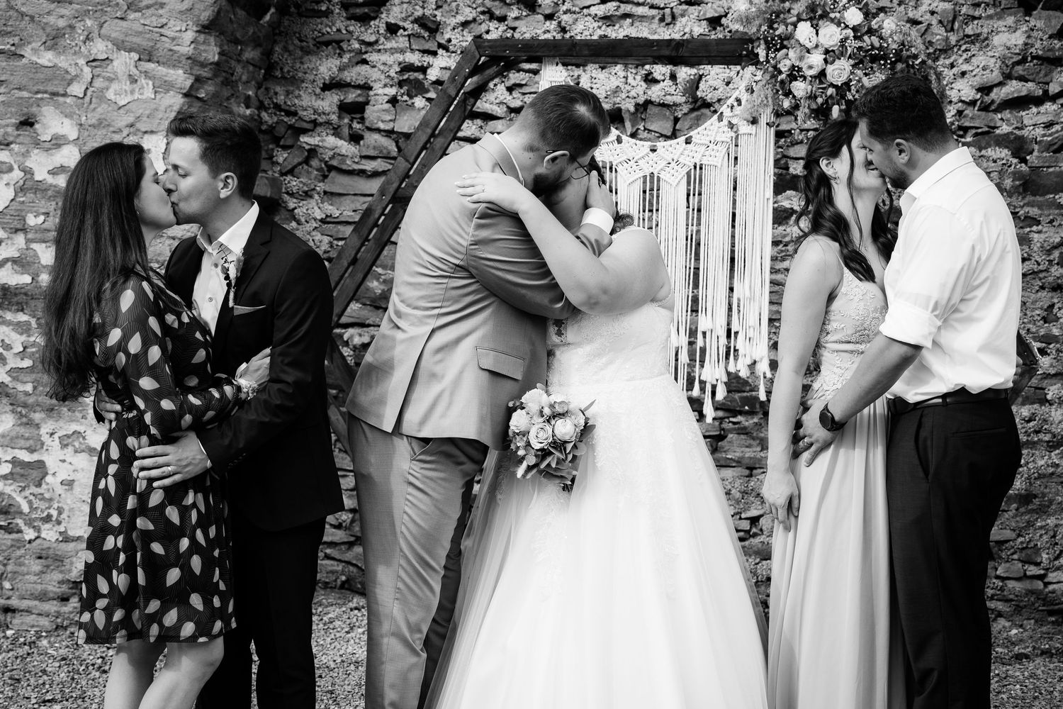Two couples share a first kiss at wedding altar with macrame backdrop in black and white photo.