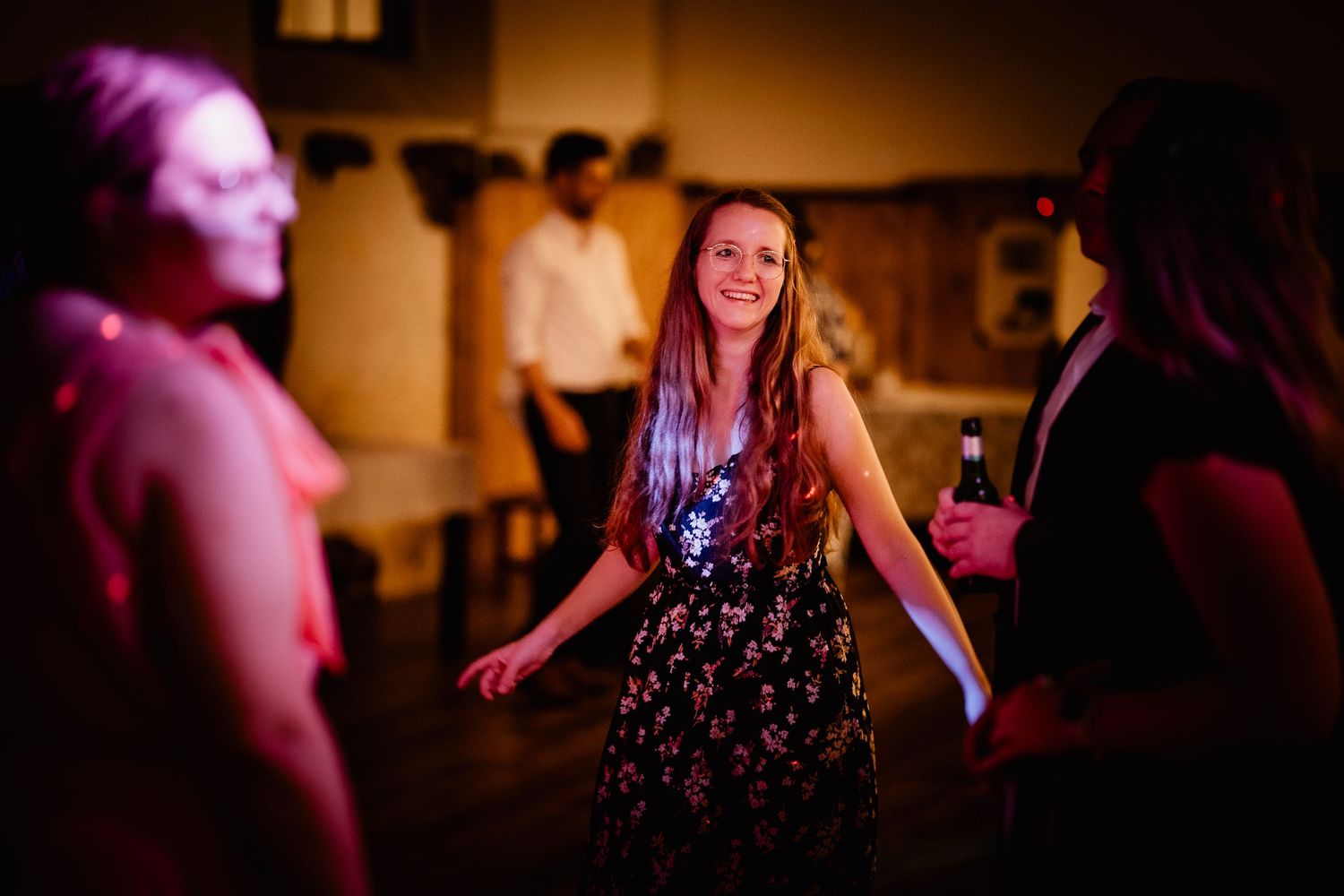 Woman in floral dress smiles and dances under warm purple lighting at lively wedding reception party.