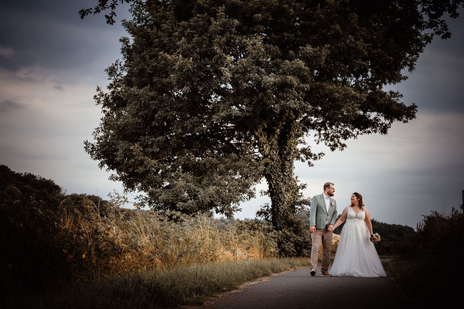 Bride and groom walking hand in hand on rural path beneath large tree at dusk.