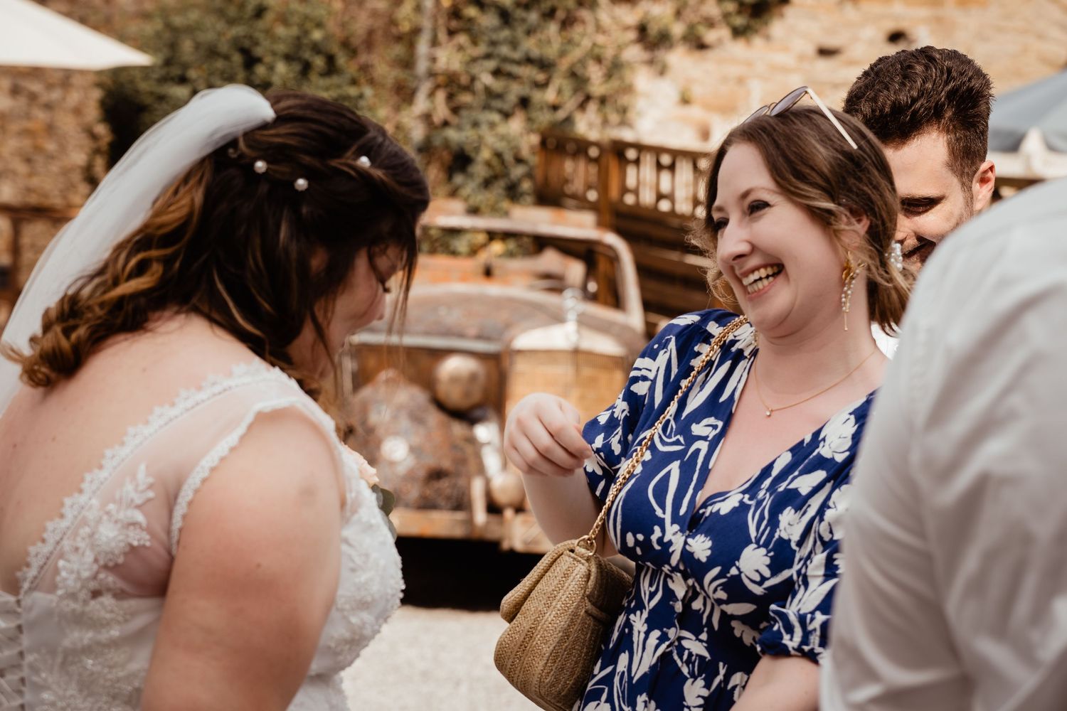 Smiling wedding guest in blue floral dress laughing with bride near vintage truck at outdoor reception.