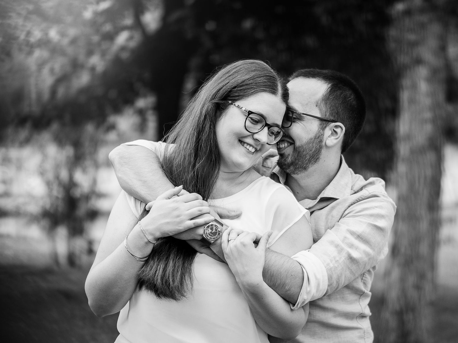Black and white photo of a couple embracing outdoors, woman smiling as man kisses her cheek.