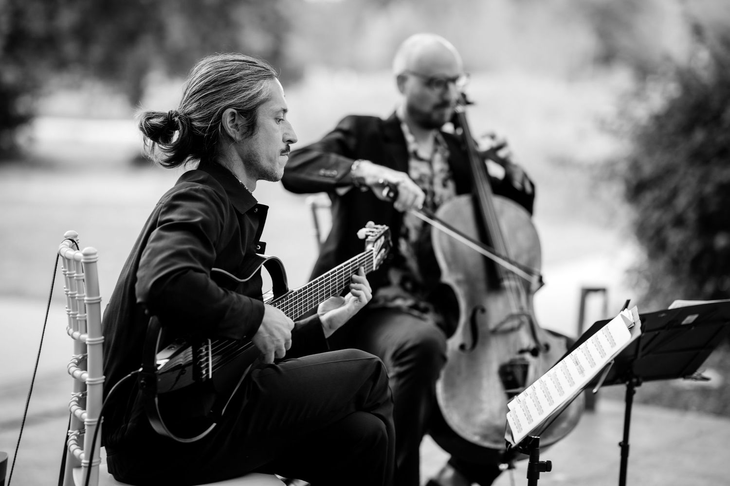 Black and white photo of guitarist and cellist performing at outdoor wedding ceremony.