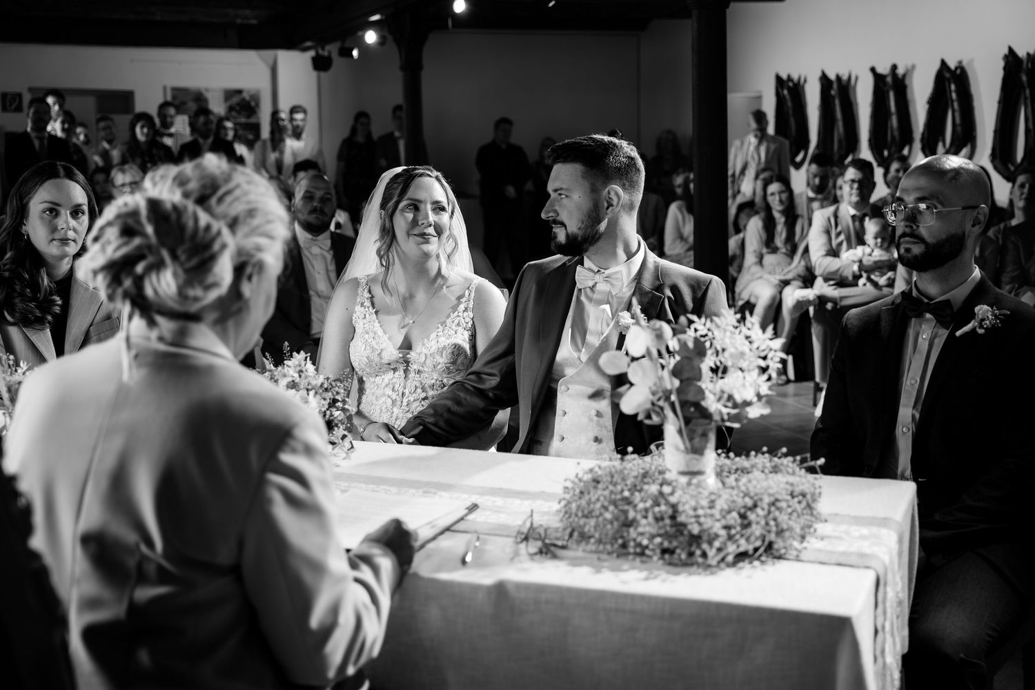 Black and white wedding ceremony photo of bride and groom at altar with guests and officiant in elegant venue.
