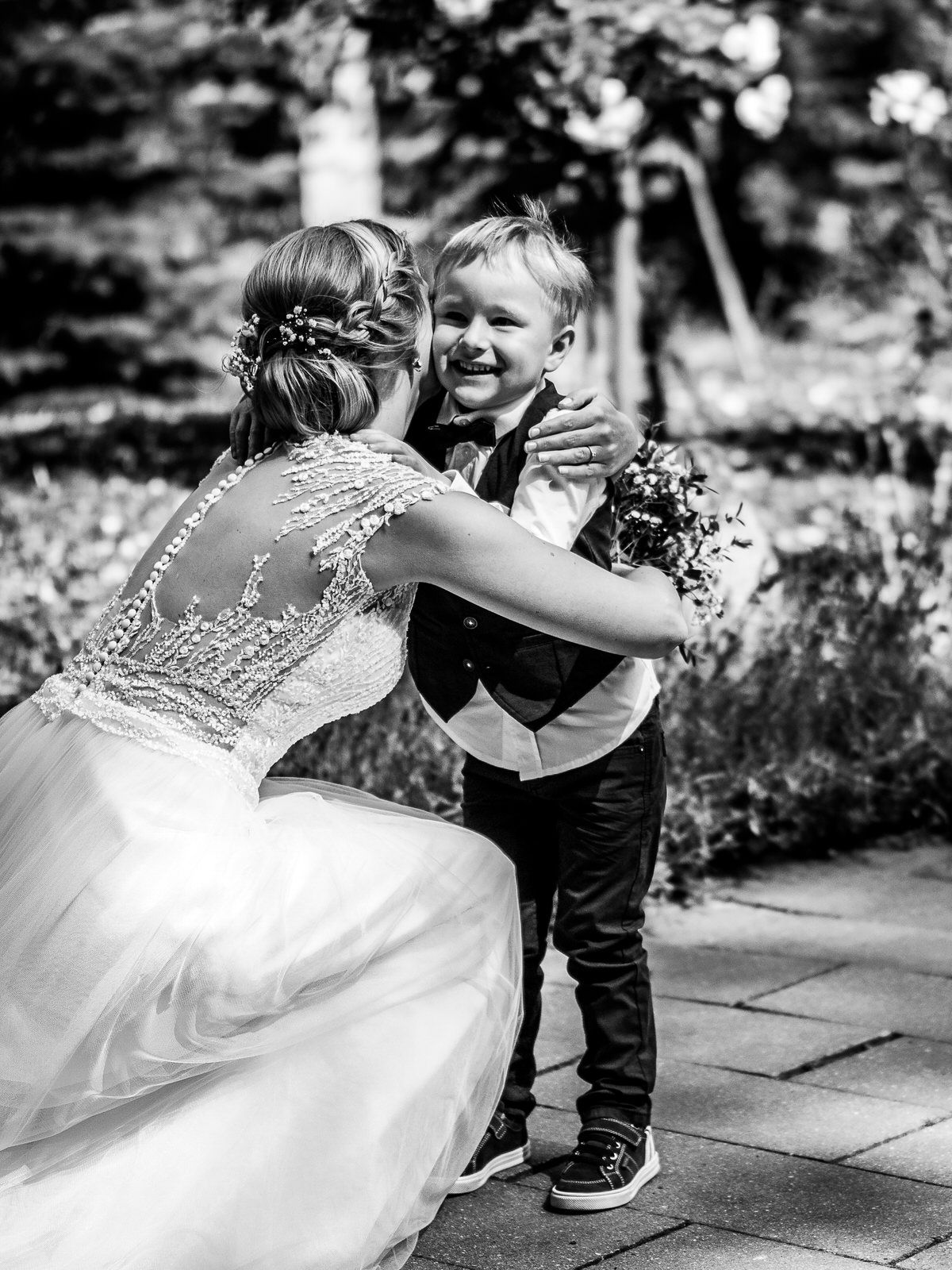 Black and white photo of a bride in lace gown hugging a smiling young boy in formal attire at a wedding.