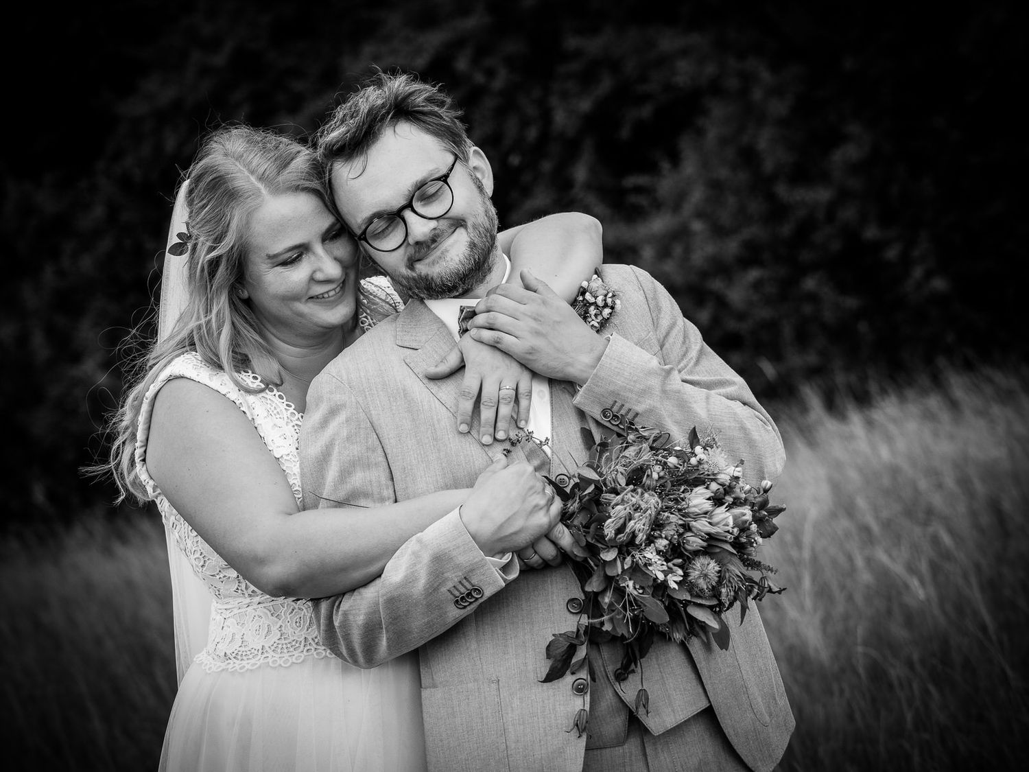 Black and white wedding photo of a smiling couple embracing outdoors, bride holding a bouquet.