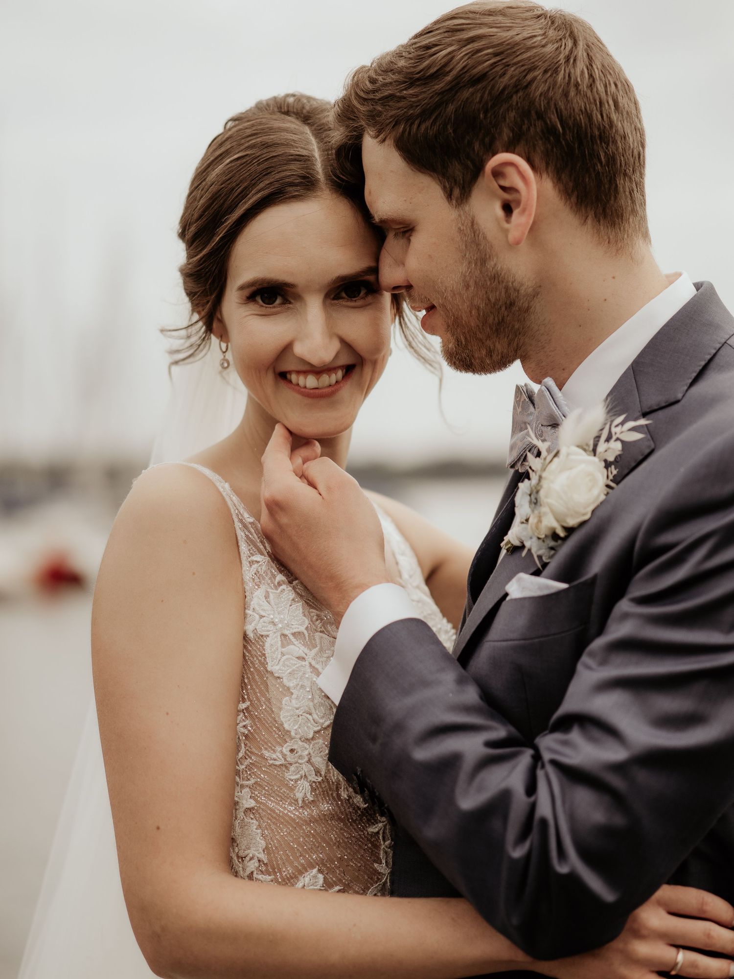 Bride and groom share an intimate moment, she smiles in lace gown as he leans close in a gray suit with boutonniere.