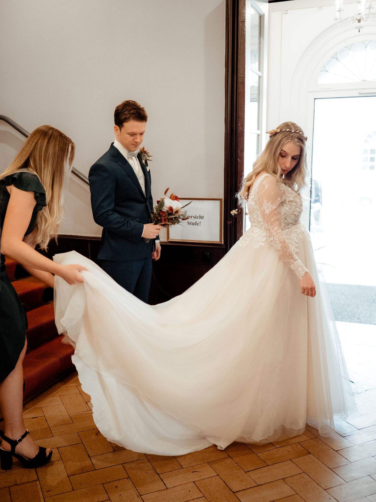 Bride in lace wedding gown has train arranged by bridesmaid while groom holds bouquet indoors.