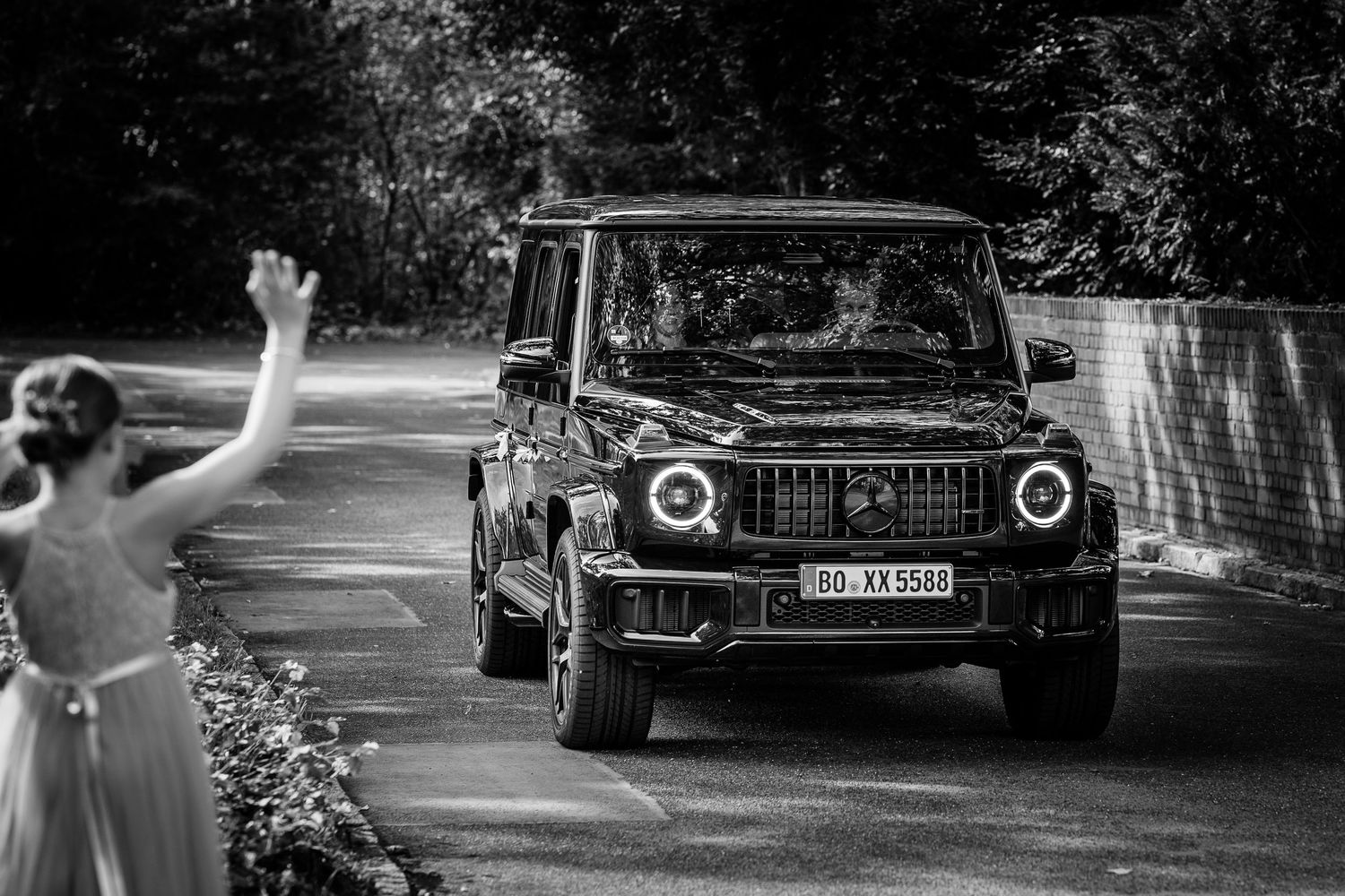 Black and white photo of a Mercedes-Benz G-Class SUV arriving at a wedding, with a bride waving in the foreground.