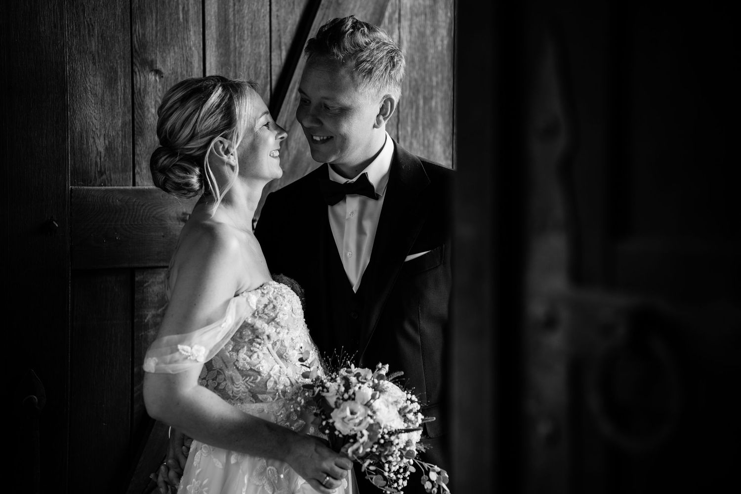 Black and white romantic portrait of bride and groom gazing at each other by wooden door.
